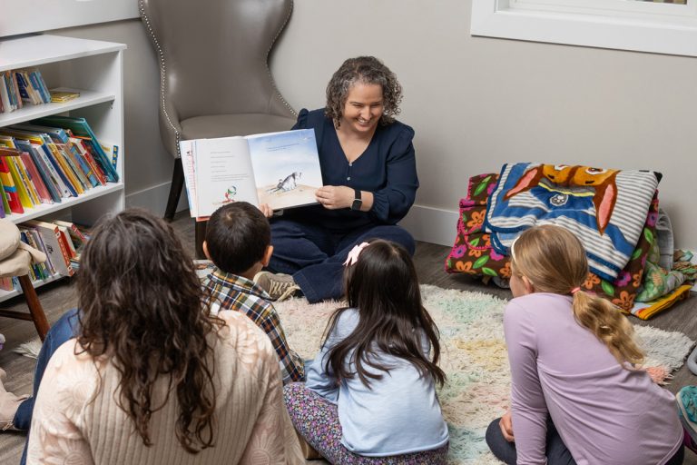 A woman reads a picture book to a group of children sitting on a rug in a cozy room with bookshelves.