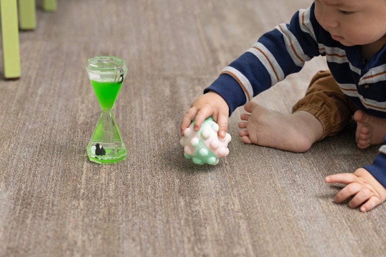 A baby plays on the floor with a textured ball near a green liquid timer.