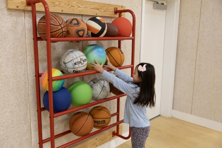 A young girl reaches for a ball on a rack filled with basketballs and playground balls in a gym.