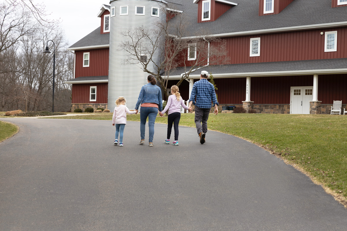 A family of four walks hand-in-hand on a driveway toward a large red barn-style building.