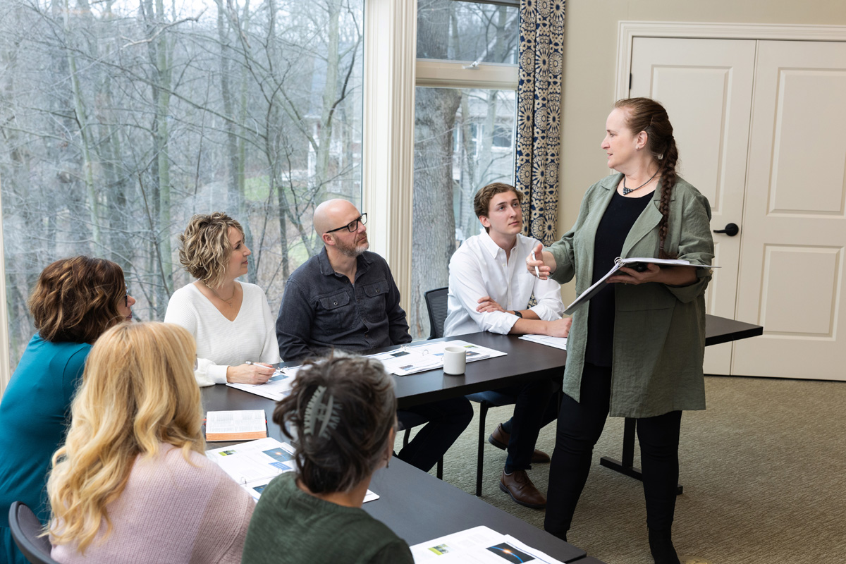 A woman stands and speaks to five seated people in a meeting room with large windows.