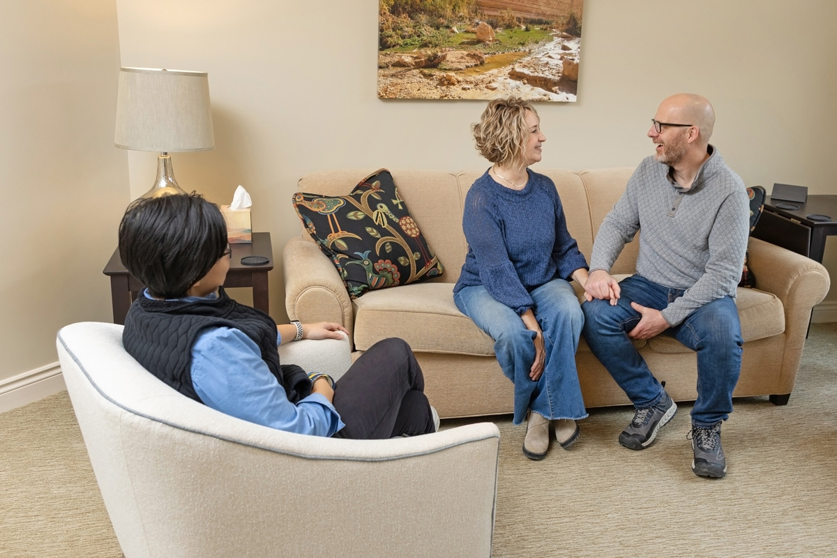 A couple sits on a couch holding hands, talking with a therapist in a cozy office setting, reflecting the warm and supportive atmosphere of a marriage counseling retreat.