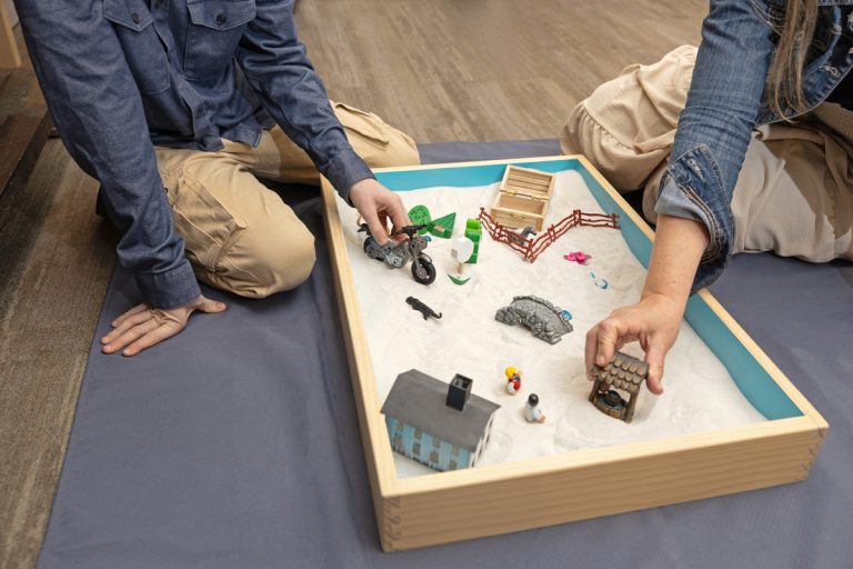Two people play with miniature toys in a sandbox on the floor, arranging figures and objects.