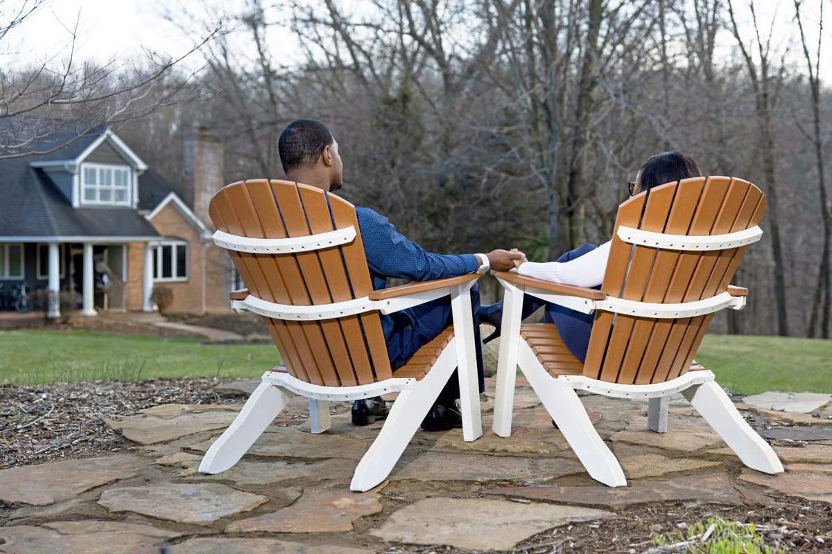 A couple sits holding hands in wooden chairs, facing a house and trees in the background.