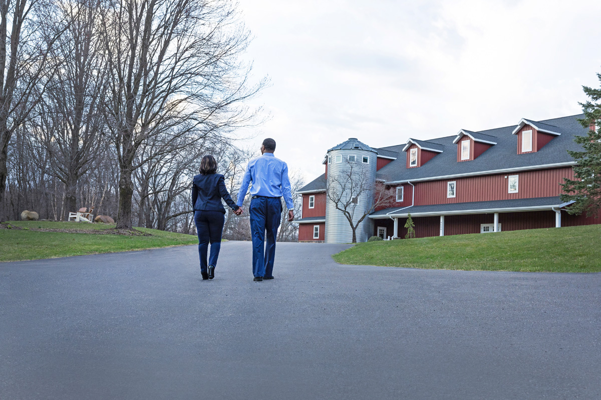 A couple holding hands walks toward a red barn on a paved path surrounded by trees and grass.