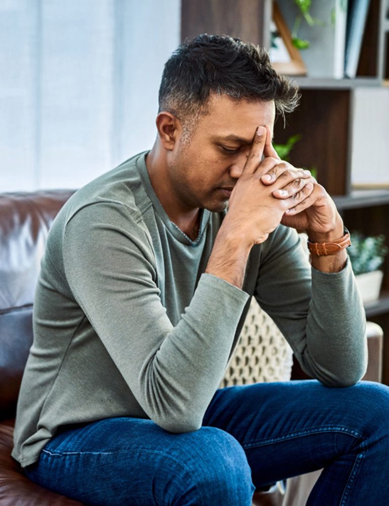 A man sits on a couch, leaning forward with his hands clasped to his forehead, looking stressed—perhaps in need of the support offered by a counseling ministry.