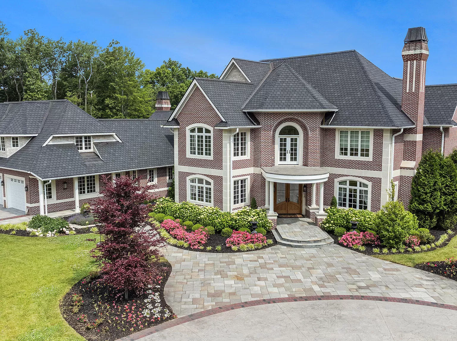Large brick house with arched windows, landscaped yard, gardens, and a circular stone driveway under a blue sky.