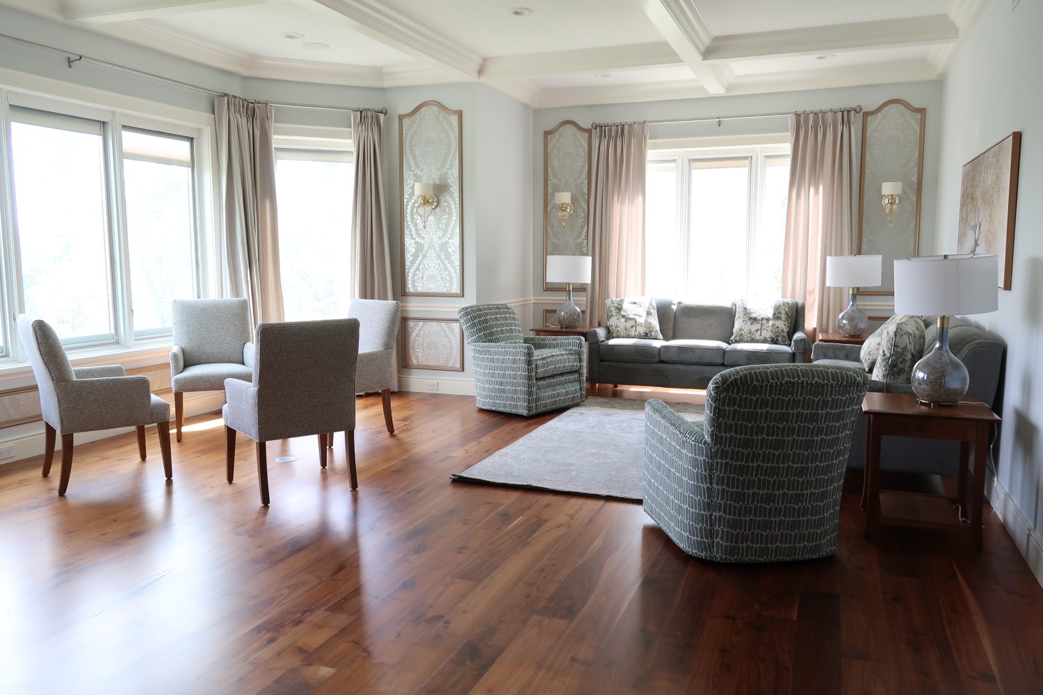 Bright living room with wood floors, gray chairs, a sofa, coffee table, lamps, and large windows with curtains.