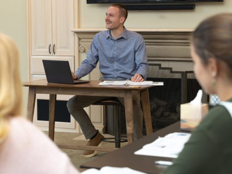 A man sits at a desk with a laptop, smiling at two people in a classroom setting.