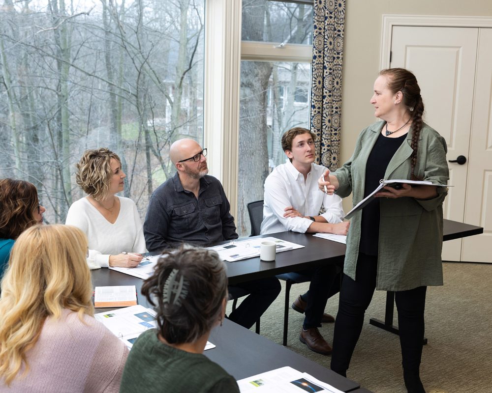 A woman stands and speaks to five seated people in a meeting room with large windows.