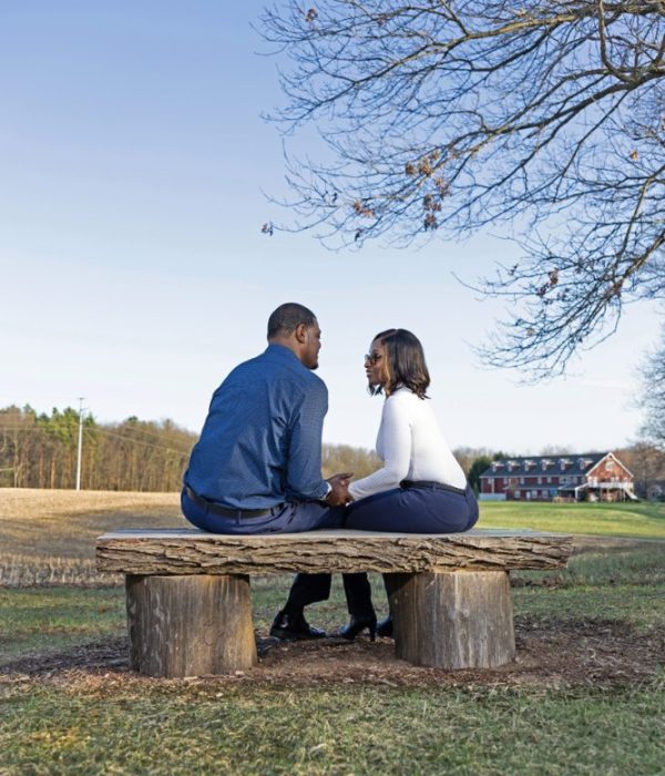 A couple sits on a wooden bench outdoors, holding hands and facing each other under a clear sky.